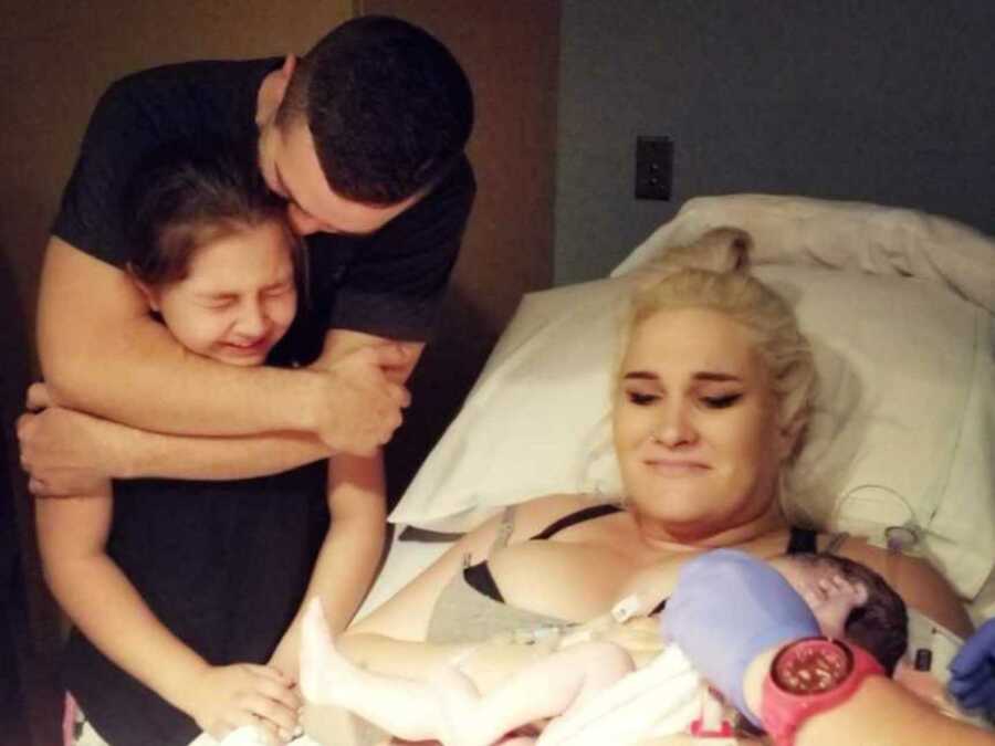 dad hugs daughter while standing next to mom and baby sister in hospital bed