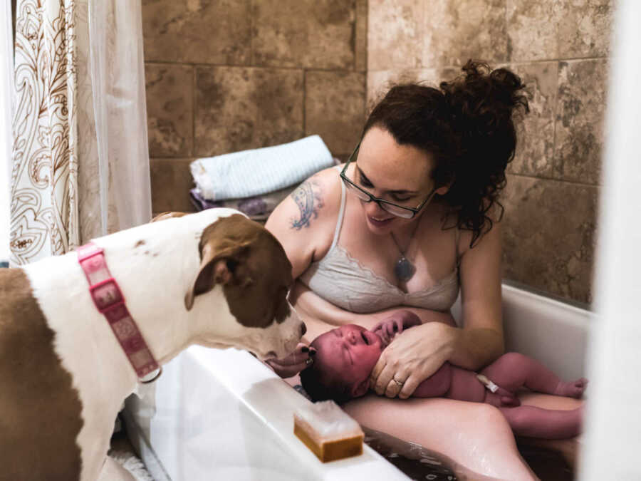 Woman who just gave birth to 11 pound baby holds newborn in lap in bathtub while dog leans over tub