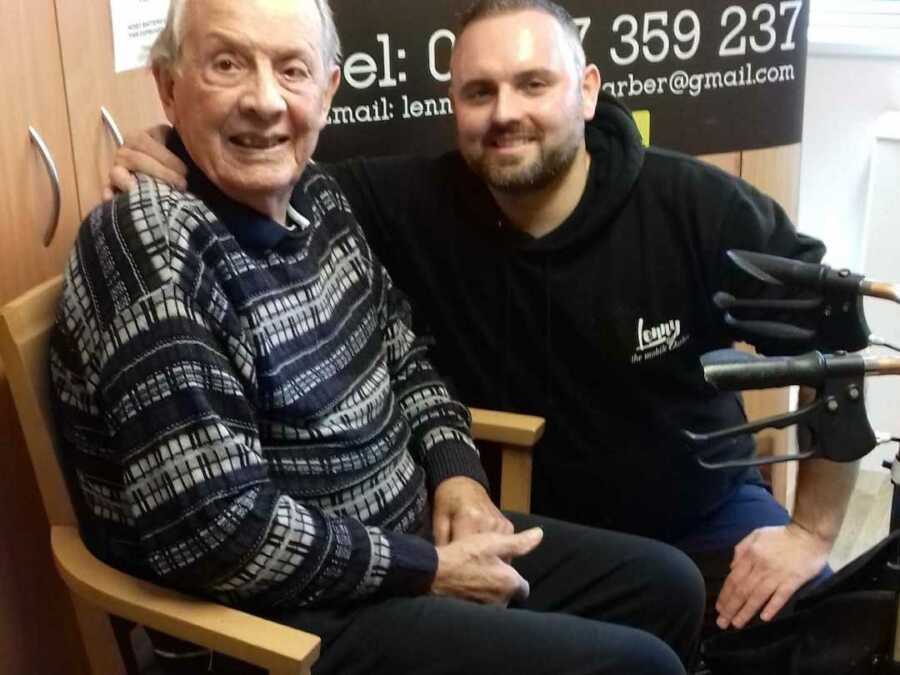 Elderly man sits in chair next to his walker while younger man crouches next to him with around him