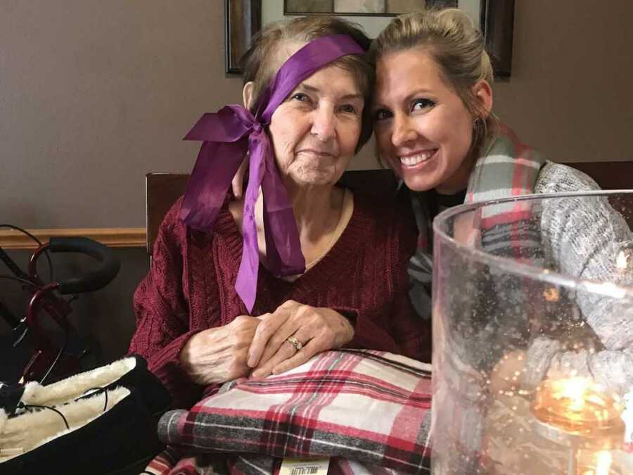 grandmother with alzheimers wears a purple ribbon with bow draped on her head sitting next to granddaughter smiling