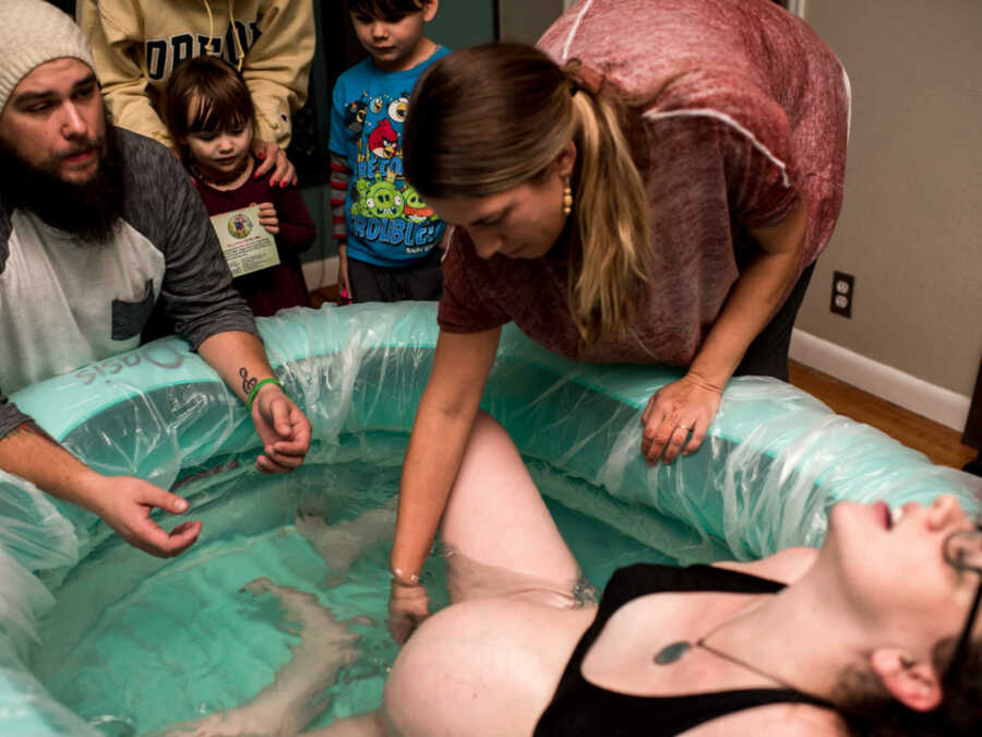 Midwife reaching into birthing pool to help woman give birth while family watches