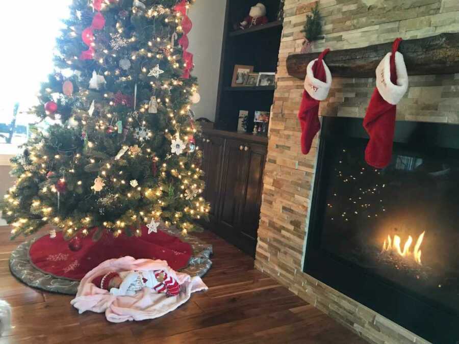 Adopted newborn babygirl lying under Christmas tree in front of lit fireplace