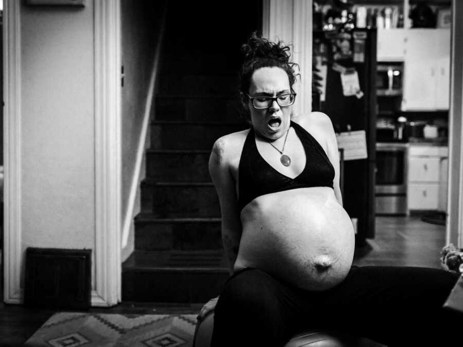 Pregnant woman sits on side of chair in home looking down at stomach in pain