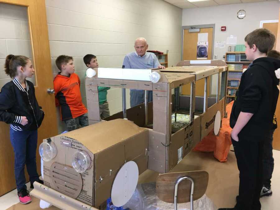 retired teacher stands with students and cardboard bus