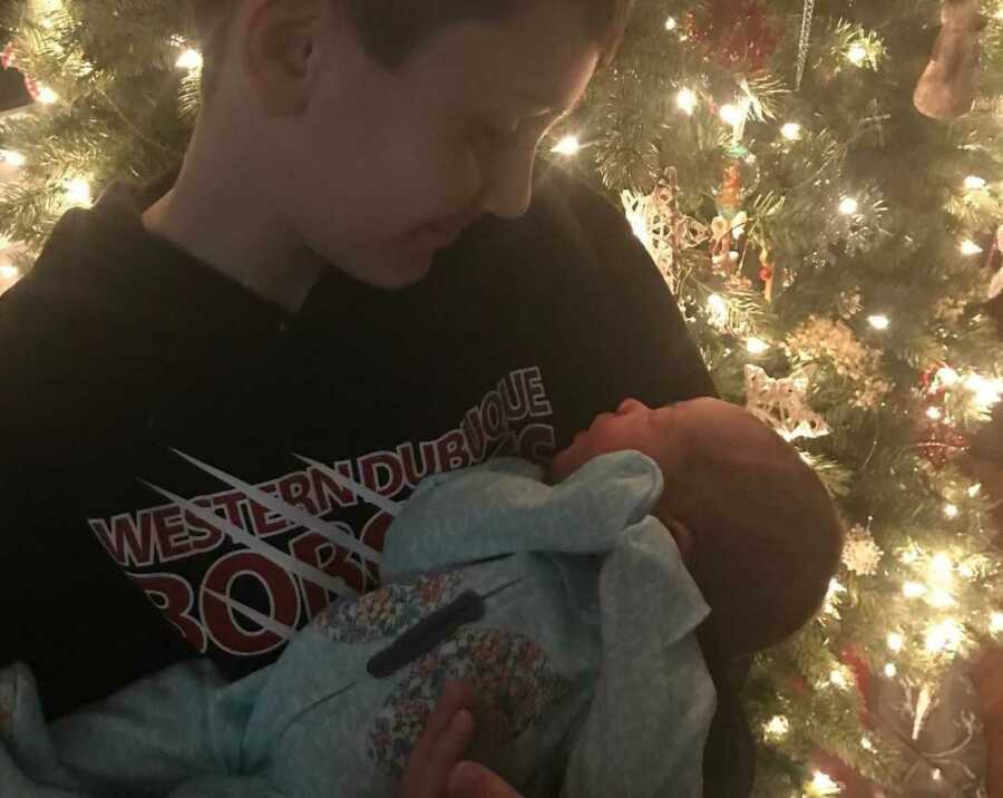 Joyous big brother holding newborn sister in front of Christmas tree