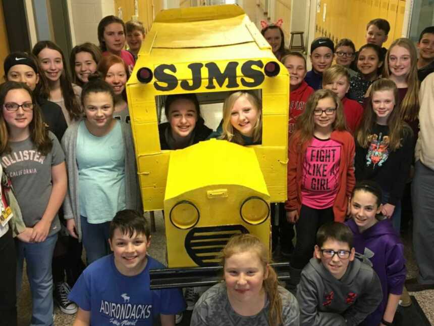 students pose with retired teacher and cardboard bus