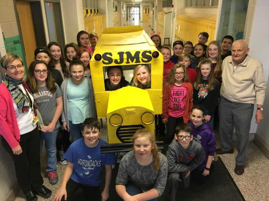 students pose with retired teacher and cardboard bus