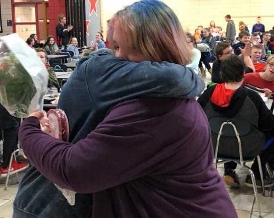 teen girl with flowers in hand hugs teen boy in cafeteria in front of class mates