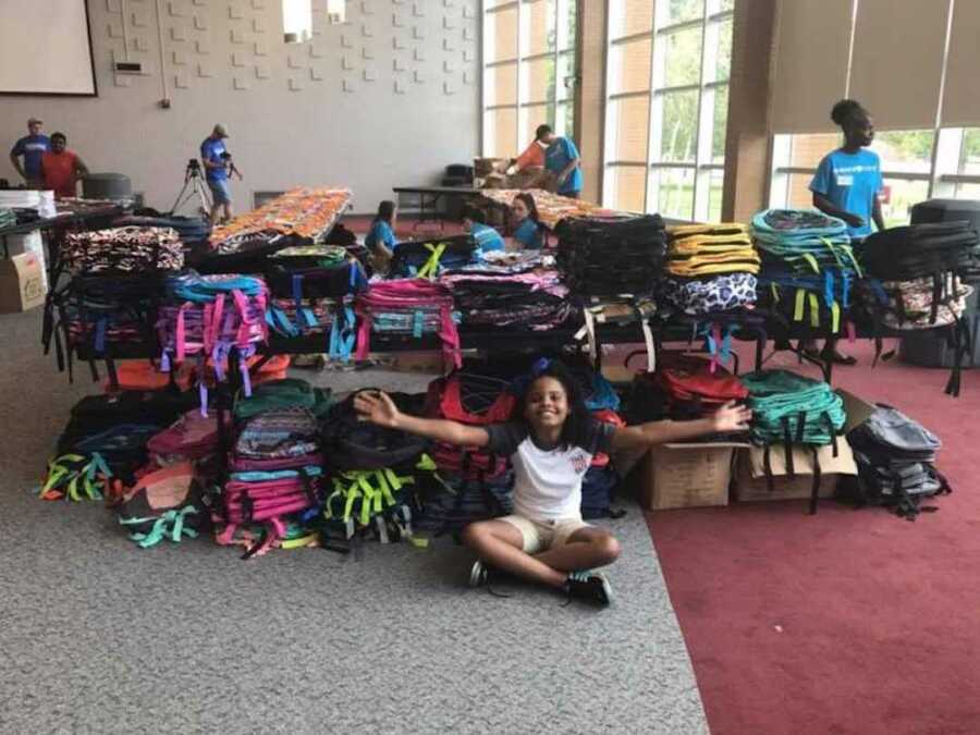 Little girl from Flint, Michigan sit on ground with arms out in front of table with stacks of backpack below and on top of it