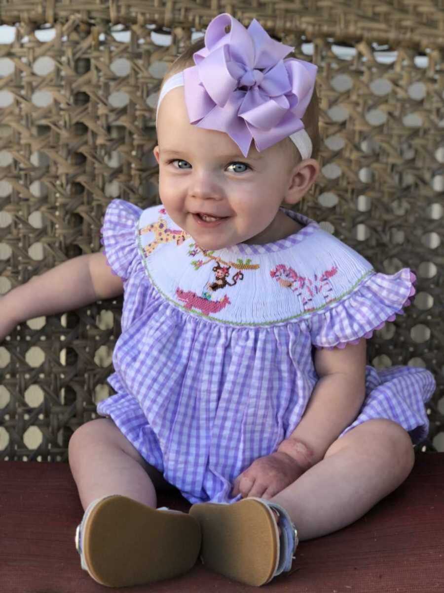 Beaming baby girl sitting on wicker chair in frilly purple outfit and bow