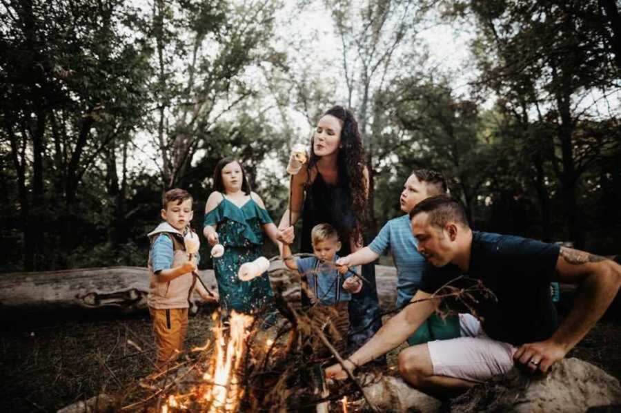 family of 6 sits by a fire outdoors roasting marshmallows 