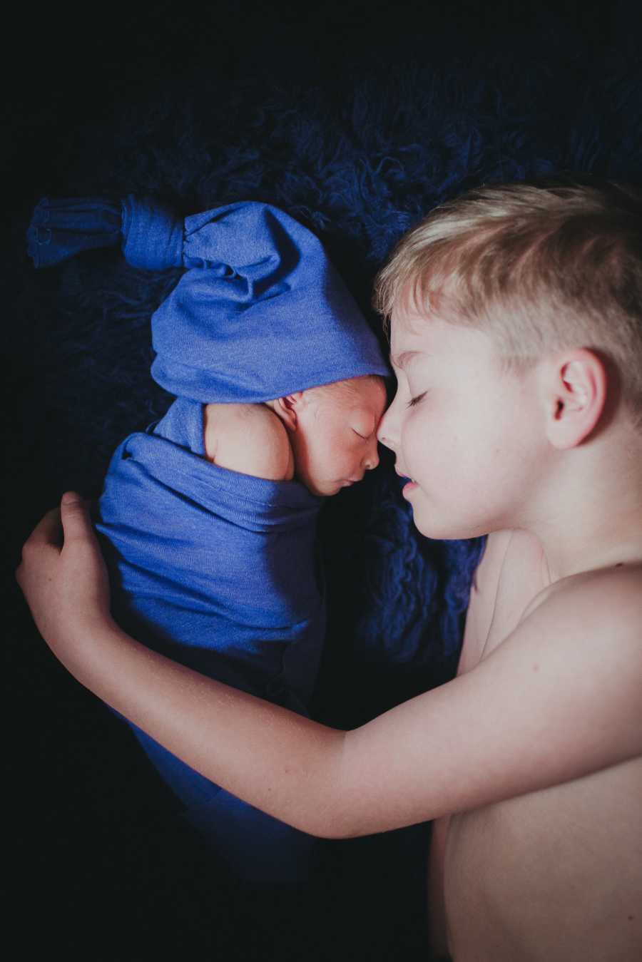 Big brother embraces his baby brother wrapped in blue blanket and hat