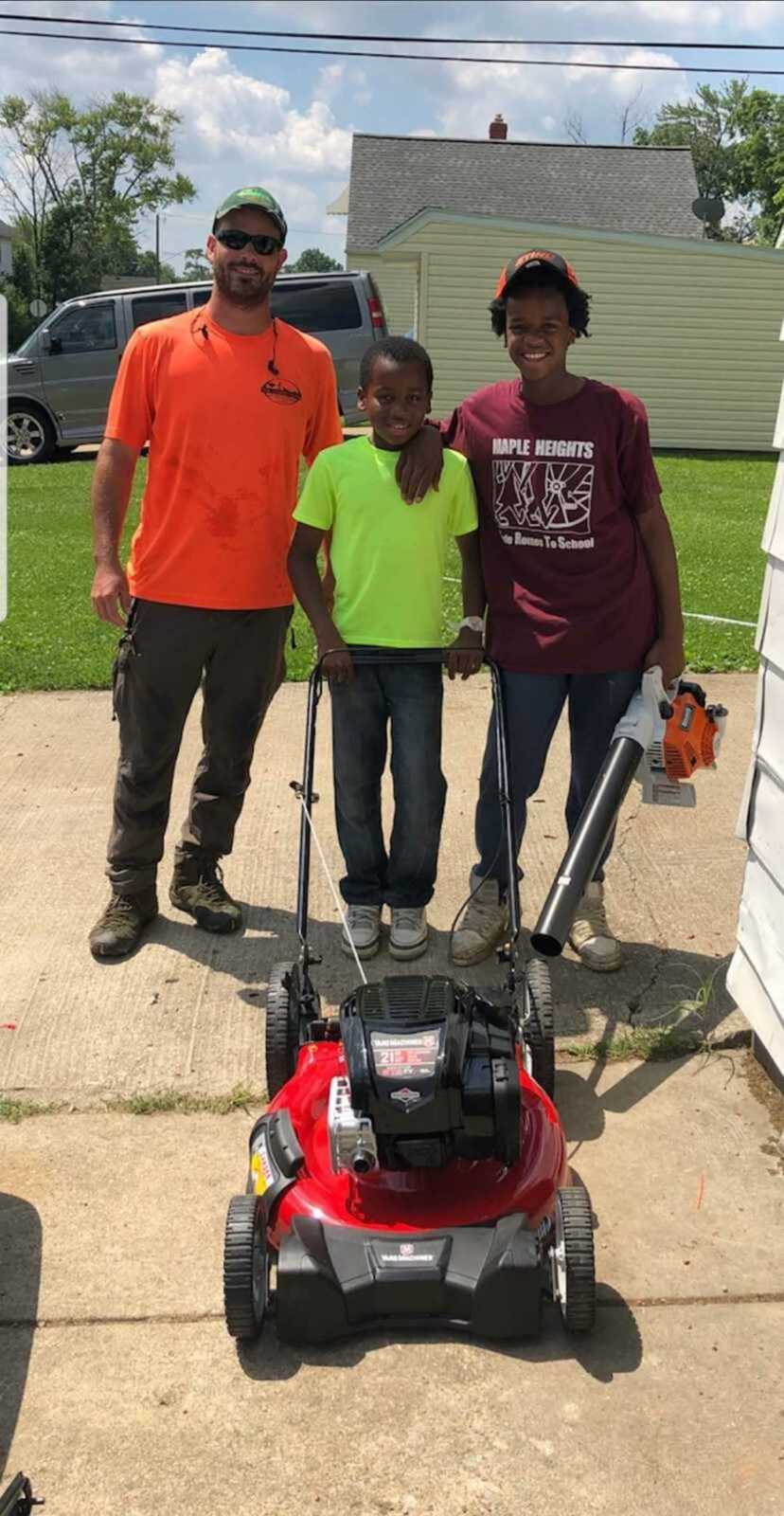 man who donated lawn mowing supplies stands with two young boys holding lawnmower and blower