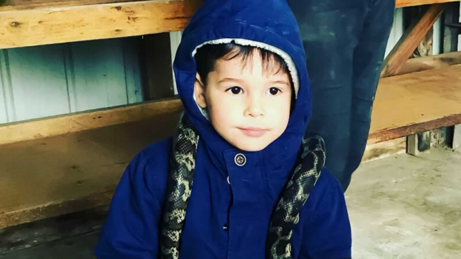 Little boy in blue coat holds a snake on his neck