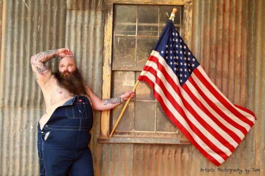 Bearded man posing in overalls holding the American flag