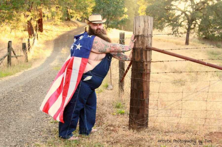 Bearded man leaning against post wearing overalls and a cowboy hat with American flag