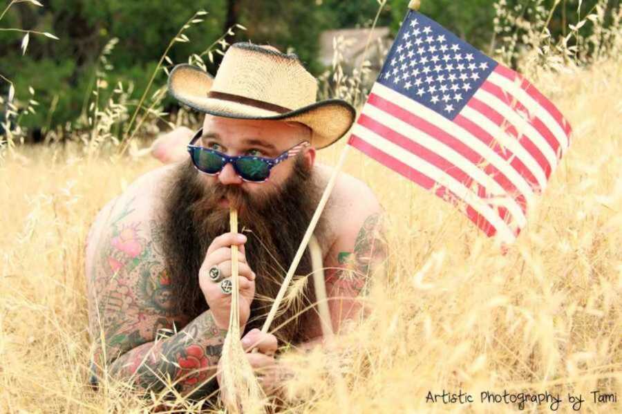 Bearded man with cowboy hat on lying in field chewing on straw holding an American flag