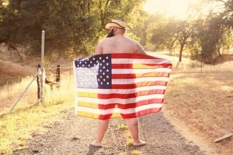 Man with beard wearing cowboy hat covering himself with an American flag on dirt road