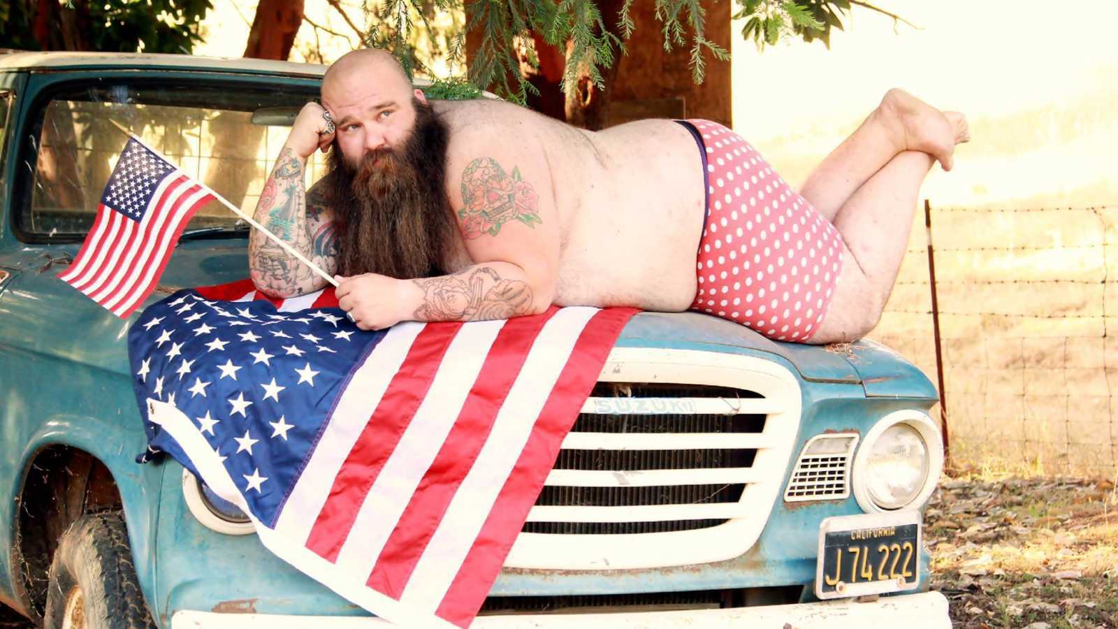 Man in beard decked out with American flags lying on top of the hood of a blue truck
