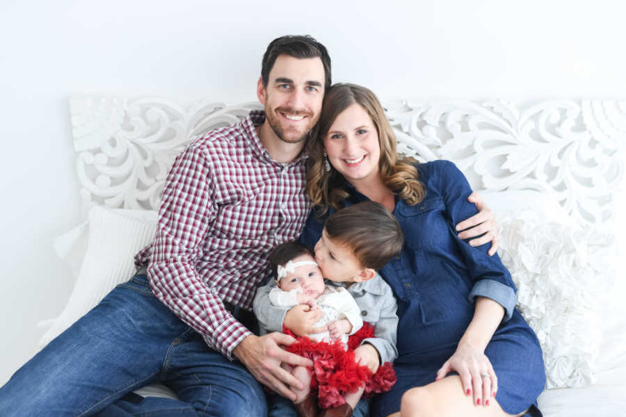 Smiling mom and dad sitting on bed embracing toddler son and newborn