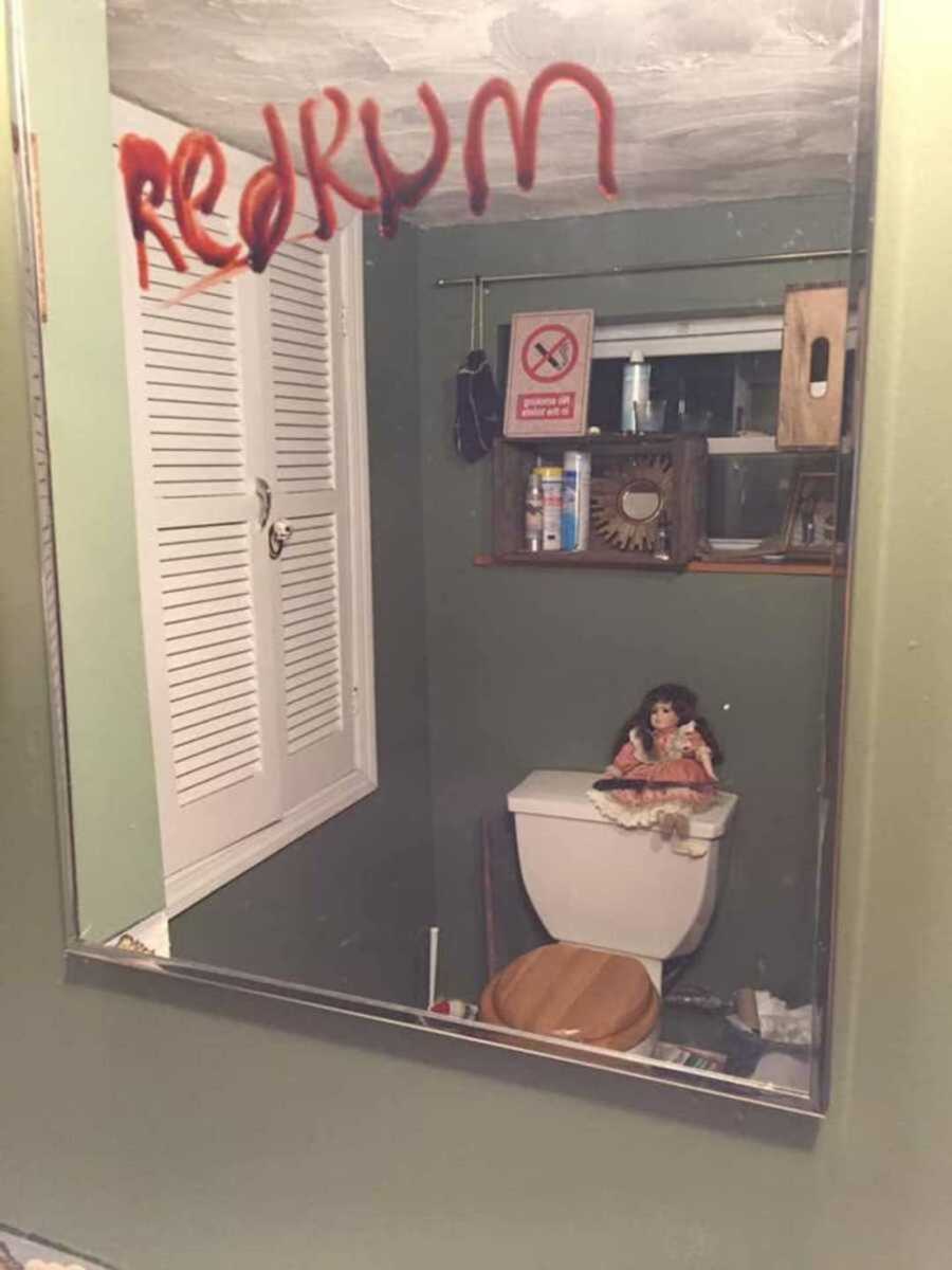 creepy doll sitting on top of toilet with knife, and "redrum" is written on the mirror