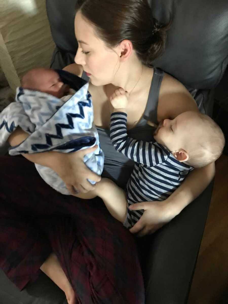 Jehovah's witness mother sits in chair with first born asleep in one arm and newborn asleep in other
