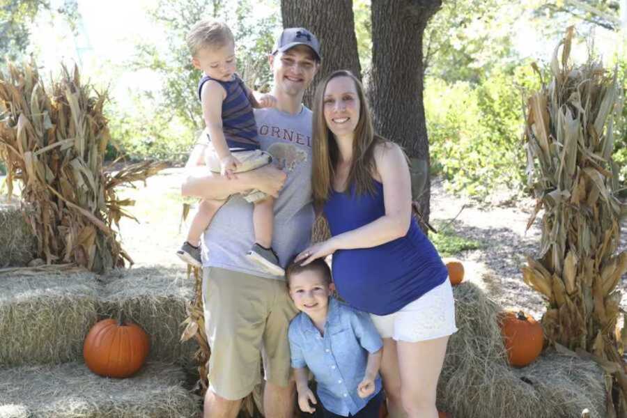 Pregnant mother stands with son who passed away beside her husband and other son in front of hay bails