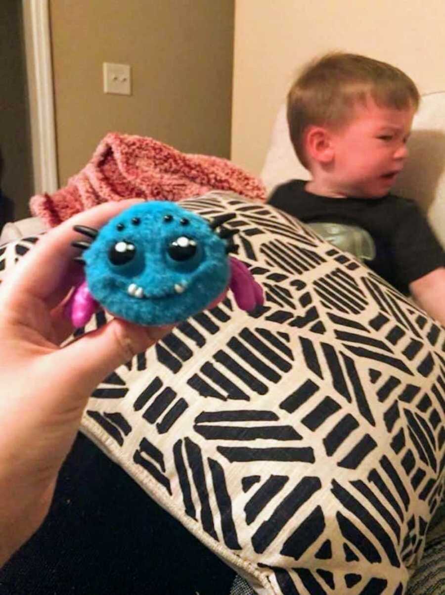 Adult hand holds fake fuzzy spider while young boy in background sits on couch crying