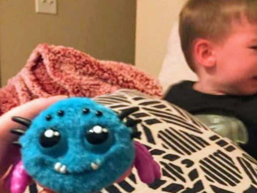 Adult hand holds fake fuzzy spider while young boy in background sits on couch crying
