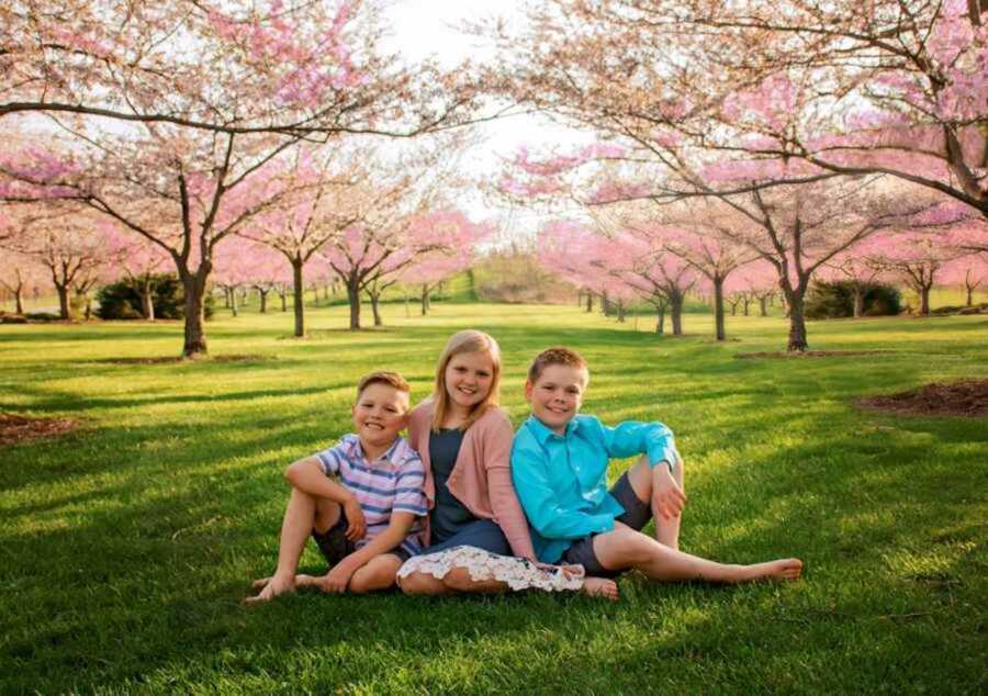 Sister and two brothers pose on grassy field next to cherry blossom trees