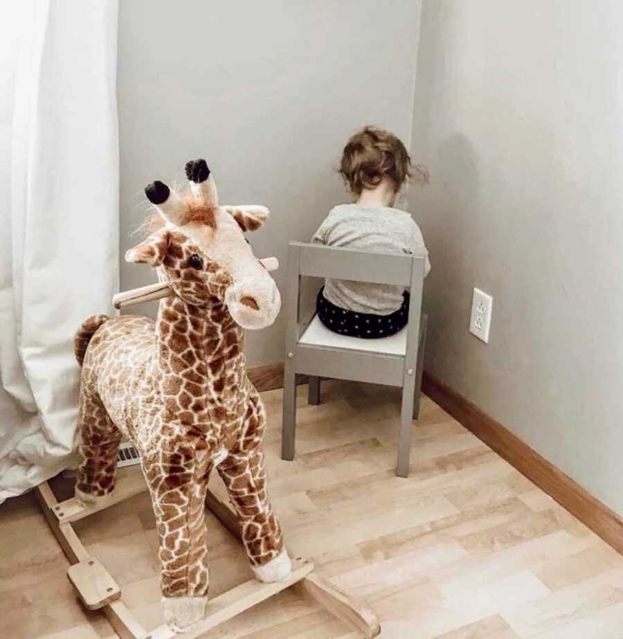 child sitting facing corner next to stuffed giraffe