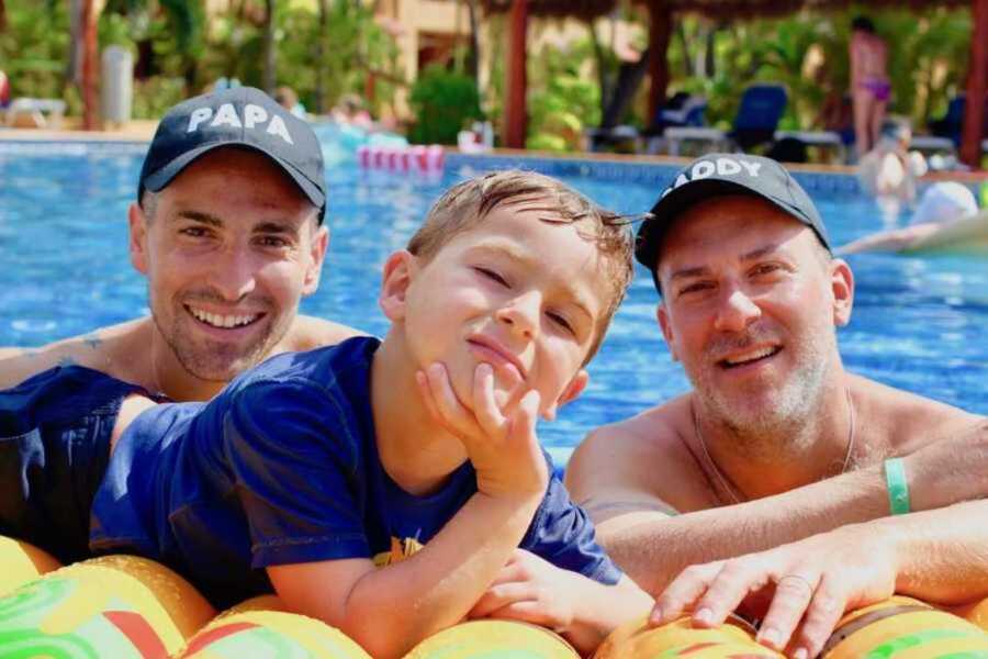 Joyous gay dads wearing hats smiling in the pool next to their son
