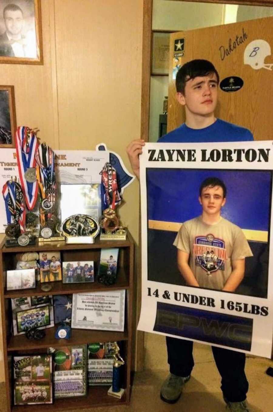 teenage boy holds wrestling banner while standing next to shelf filled with sports accomplishments