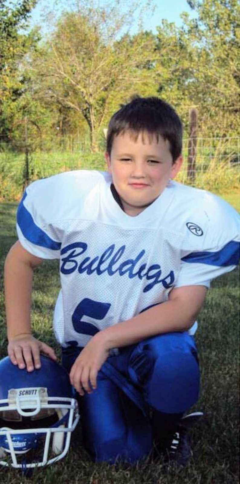 young boy kneels next to football helmet wearing uniform