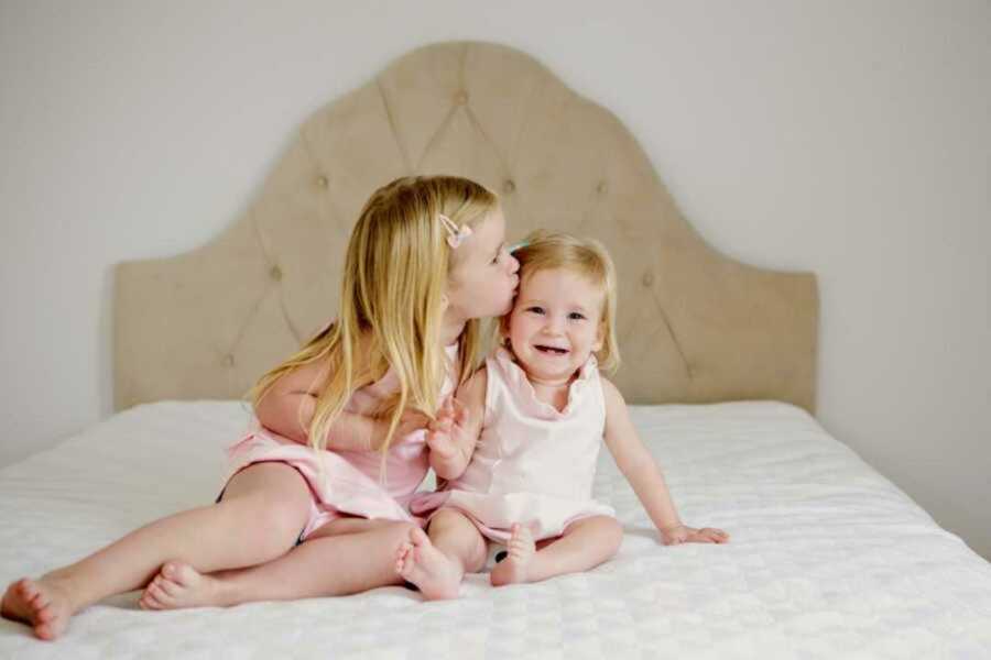 Big sister kissing smiling little sister on head on a white bed in matching pink outfits
