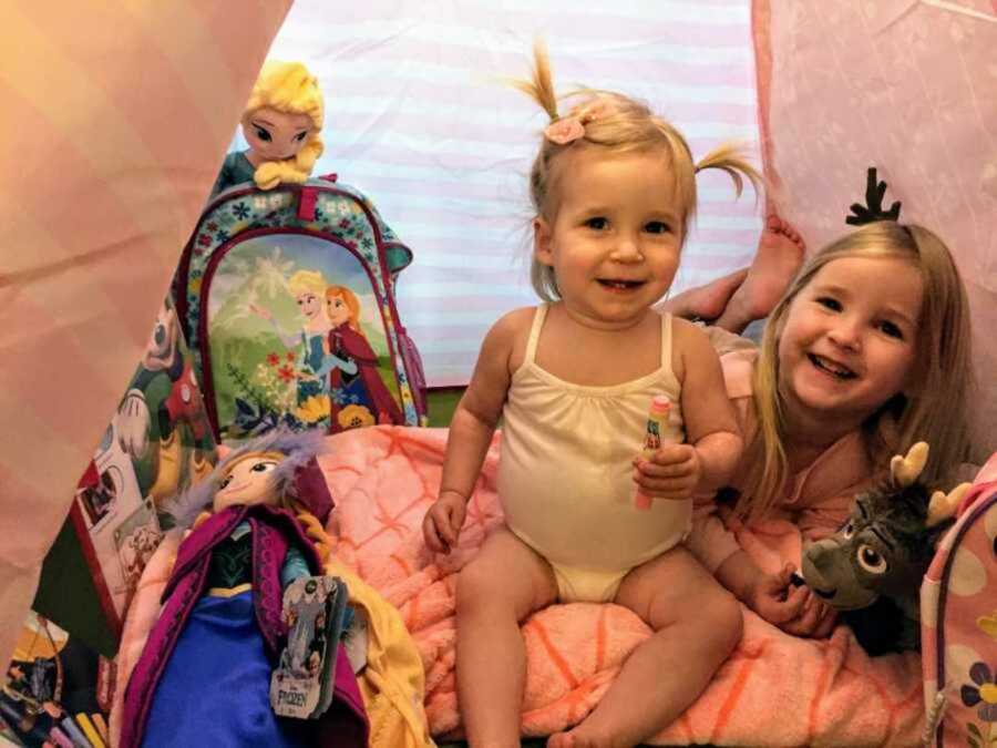 Cheerful sisters sitting in tent surrounded by colorful Disney toys