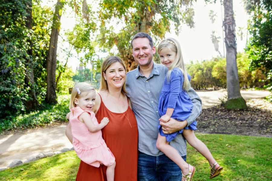 Grinning mom and dad holding their two happy daughters in bright green woods