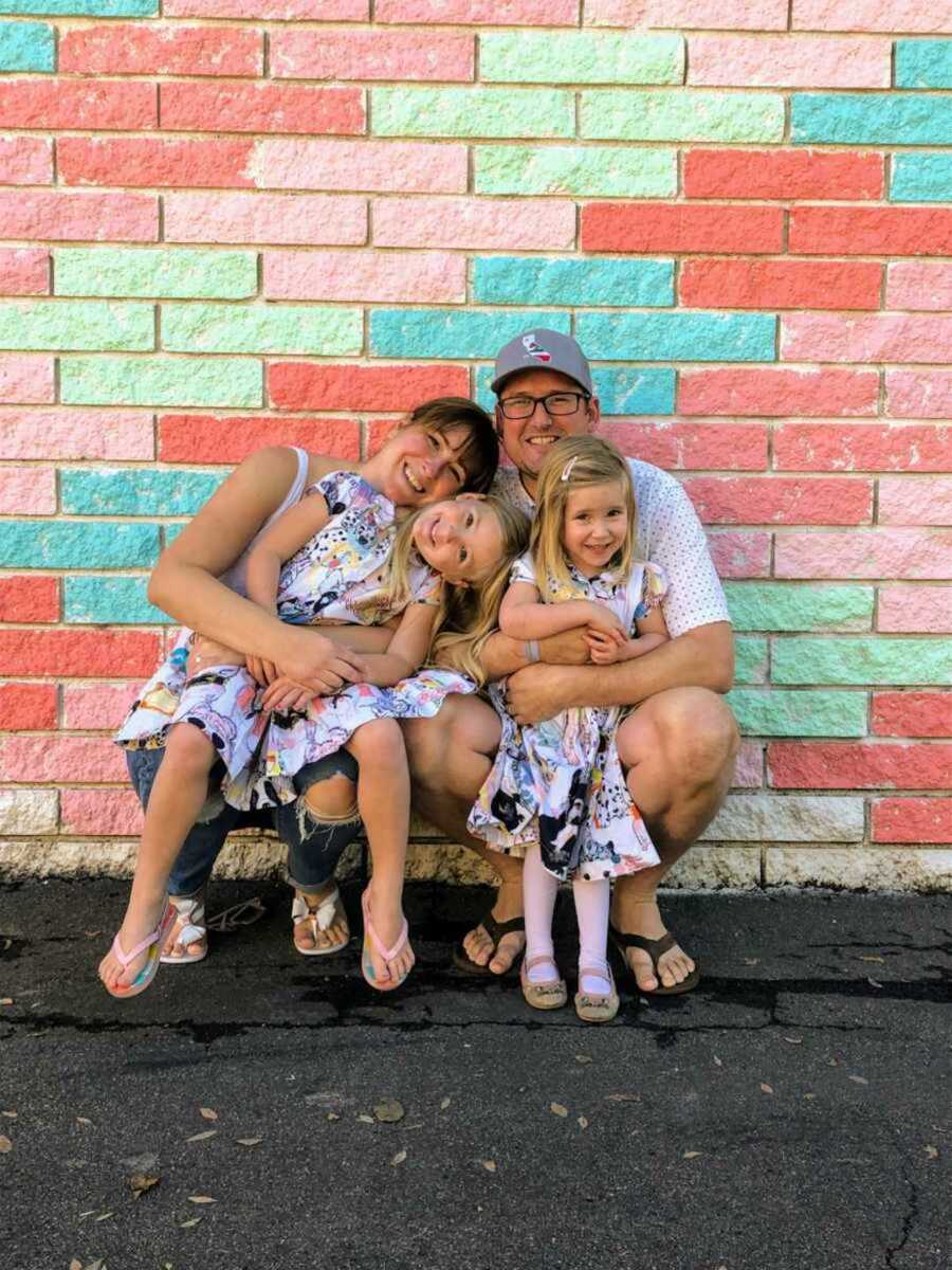 Cheerful mom and dad holding daughters in colorful dresses against a bright brick wall