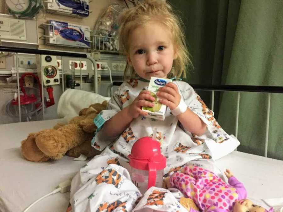 Sick toddler in hospital bed and gown sipping on a juice surrounded by toys