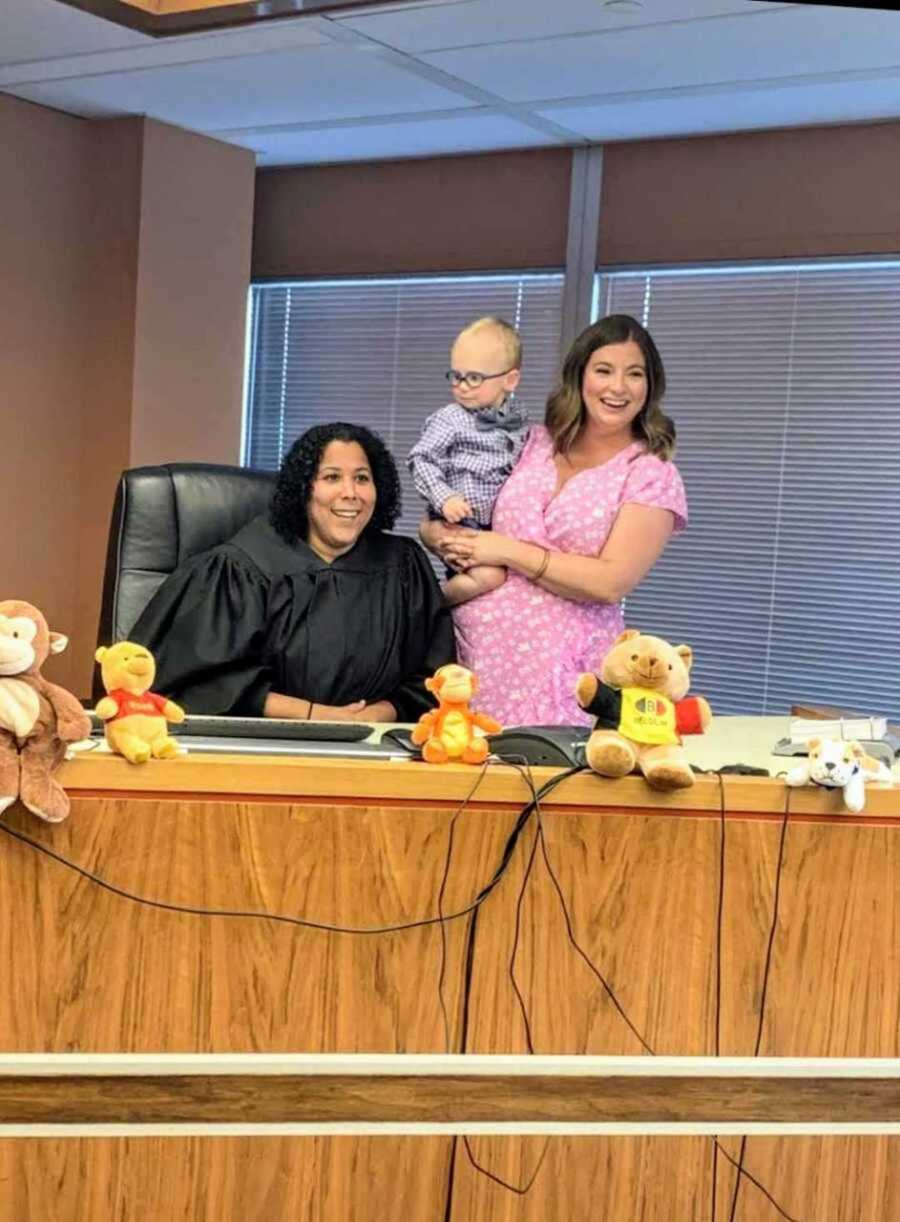 Proud foster mom holding adopted little boy in courtroom next to judge