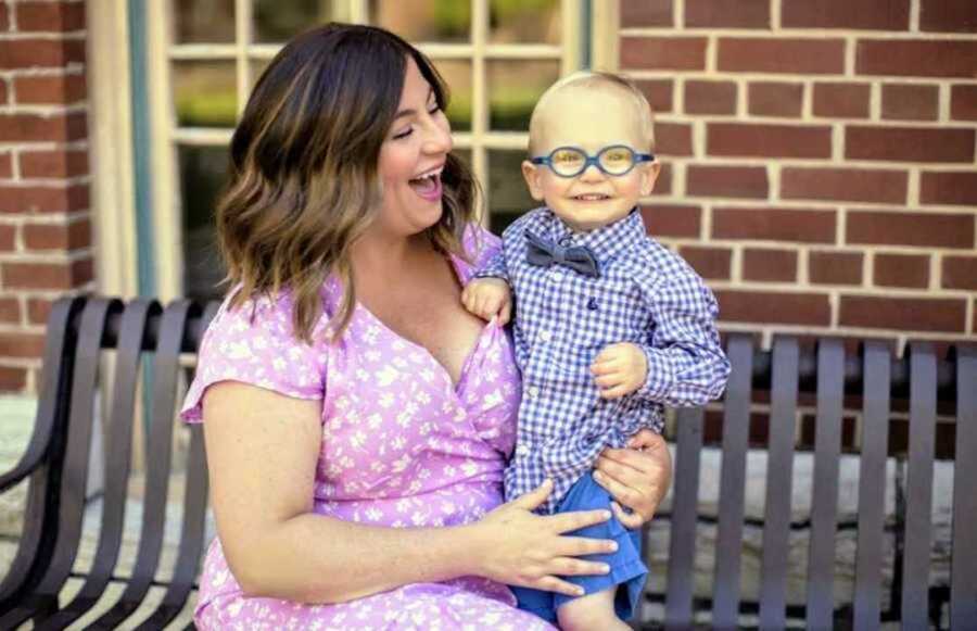 Smiling foster mom in pink dress with newly adopted son in glasses and bowtie sitting on bench