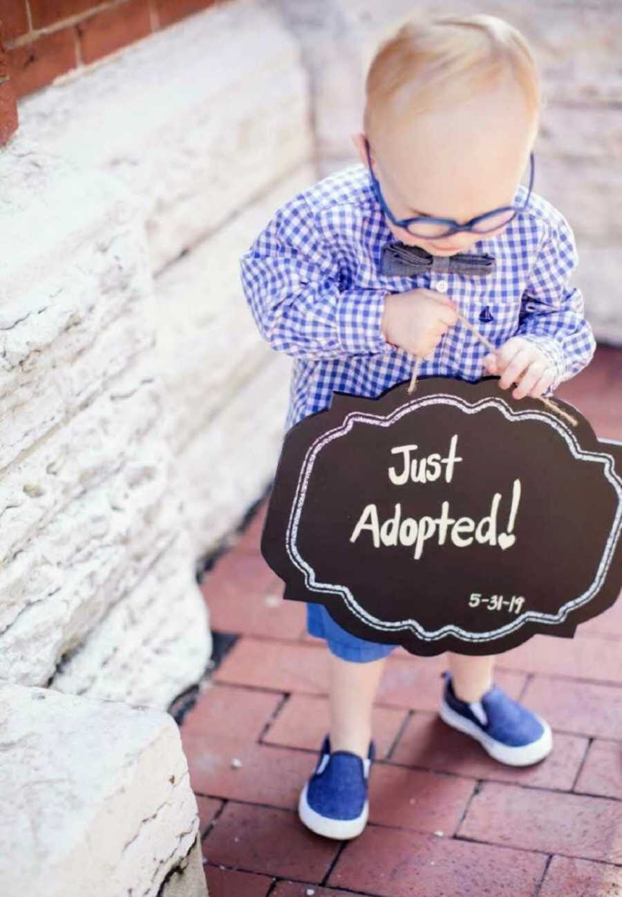 Newly adopted little boy in glasses and bowtie holding up adoption sign