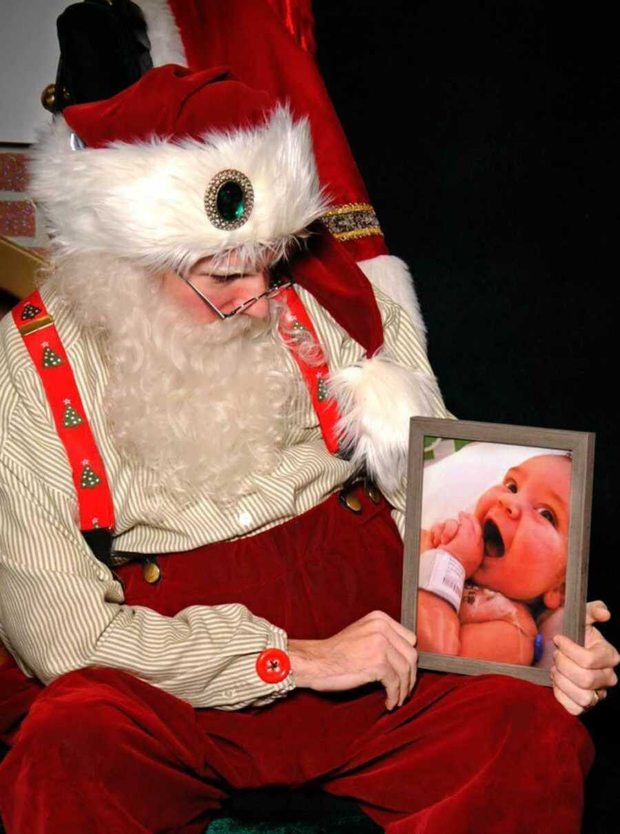 Santa Clause in red outfit holding a photo of a baby with hospital wristband on