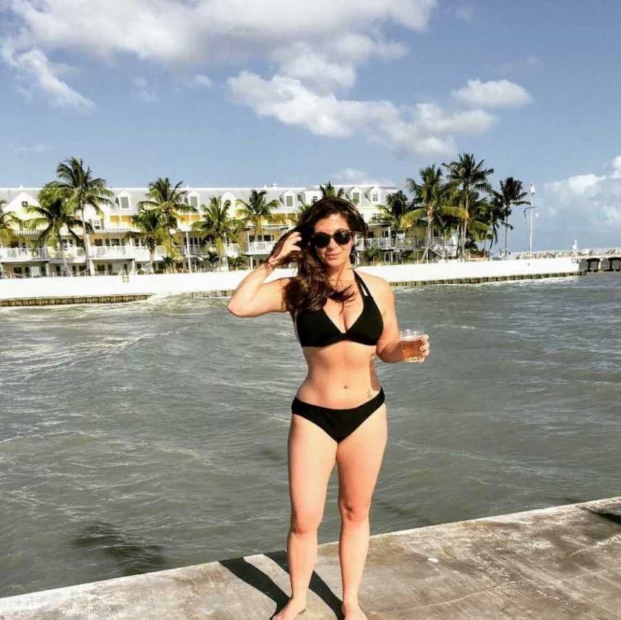 Single woman smiling in black bikini and sunglasses holding a drink by the water