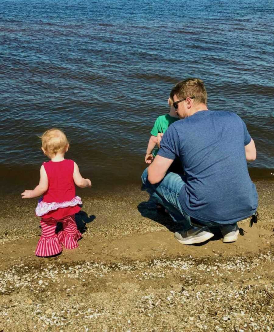 Father crouches by the shore of a lake with his young son and daughter