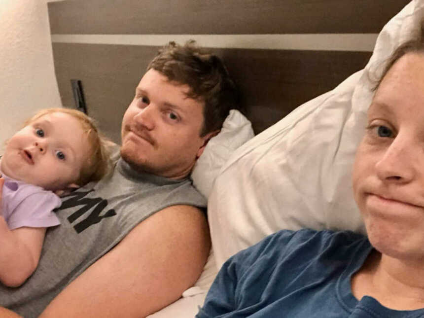 husband and wife lay in hotel room bed, husband has daughter on chest