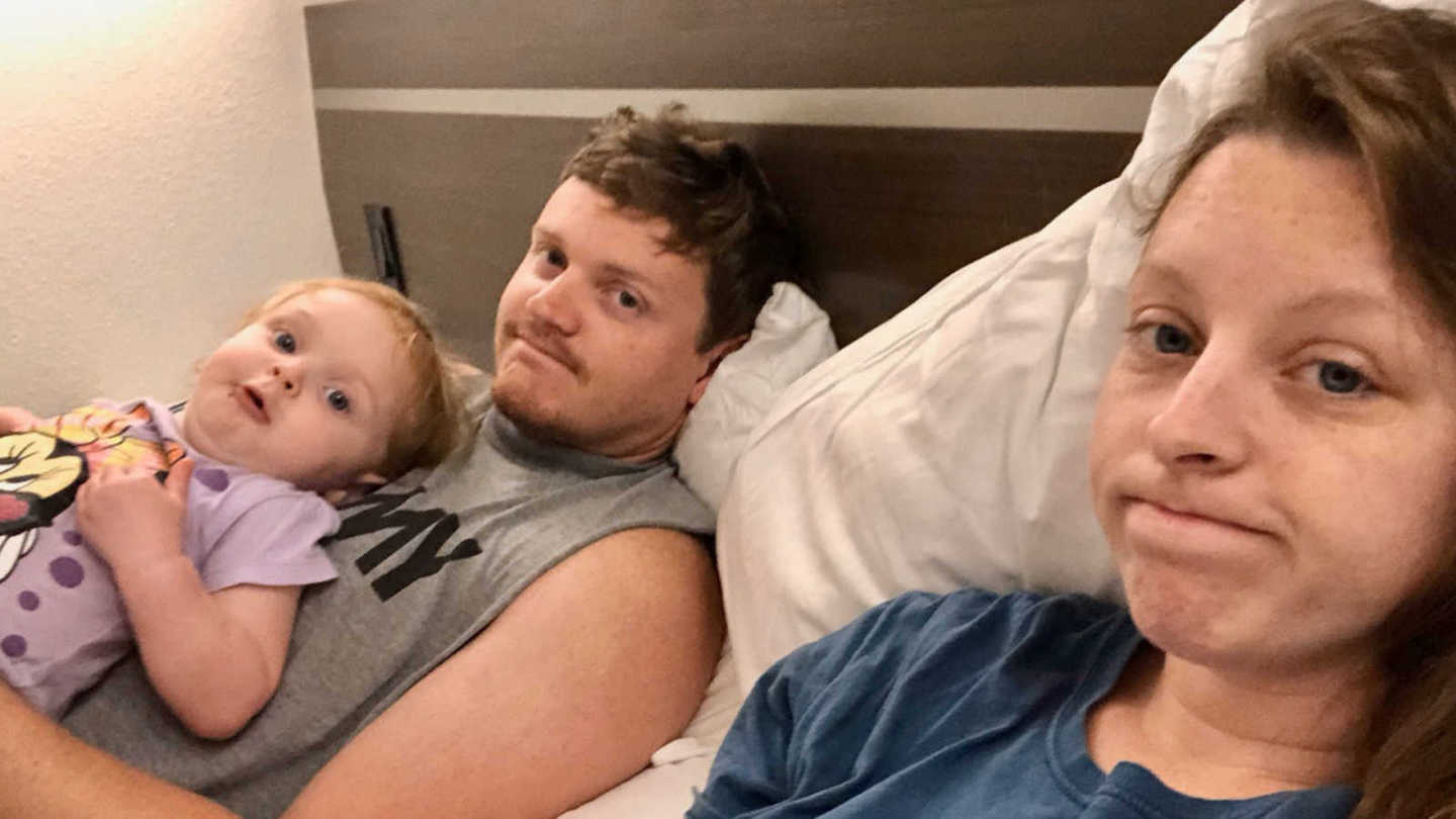 husband and wife lay in hotel room bed, husband has daughter on chest