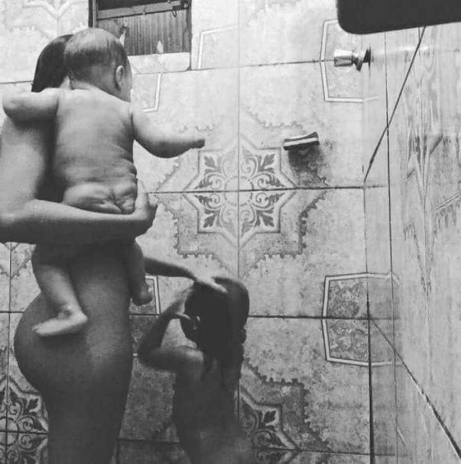 Mother stands in shower with young daughter while holding infant