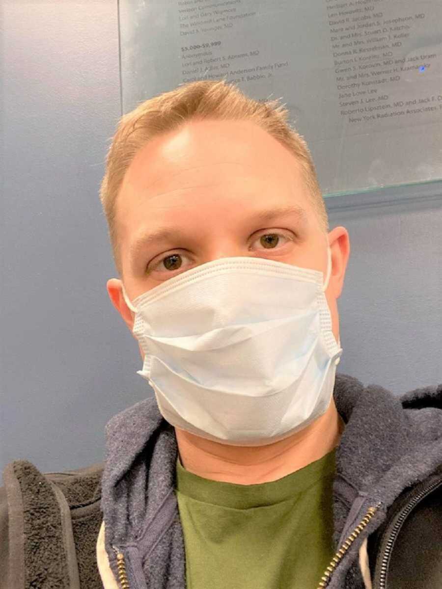 Brown eyed man with mask on wearing a green shirt in front of blue wall