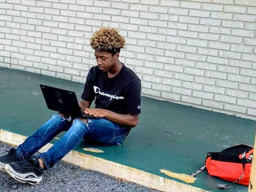 boy sits on curb outside of Subway with laptop completing schoolwork